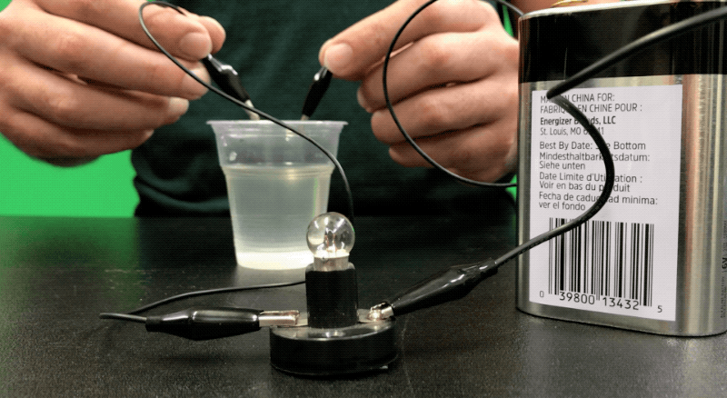 A person is seen holding two wires, one in each hand, and they are putting the wires into a clear plastic cup filled with water.