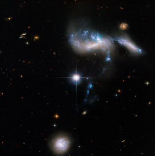 The stars, gas, and dust seem to form a blue, white, and purple knot-shaped bow above-right of center, with a small red-orange spiral in the background in the upper right. A bright white foreground star just left of center. Another smaller, purple-white spiral at the bottom center.
