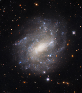 A sparkling galaxy with a bright, glowing core and patchy spiral arms dotted with pale blue star formation.