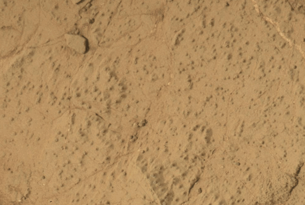 This animated image shows a close-up view of a flat, dusty, tan-colored rock surface with fine cracks and small, dark specks scattered across it. In the animation, a hole appears in the center of the frame, with loose powdered material surrounding the hole. The surrounding rock surface remains unchanged, highlighting the freshly drilled hole and the fine texture of the rock.