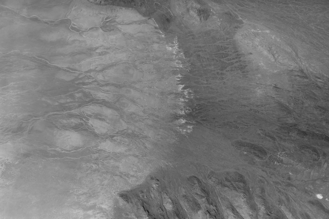 Grayscale aerial image of rugged desert terrain showing branching channels, ridges, and broad textured plains with contrasting light and dark tones.
