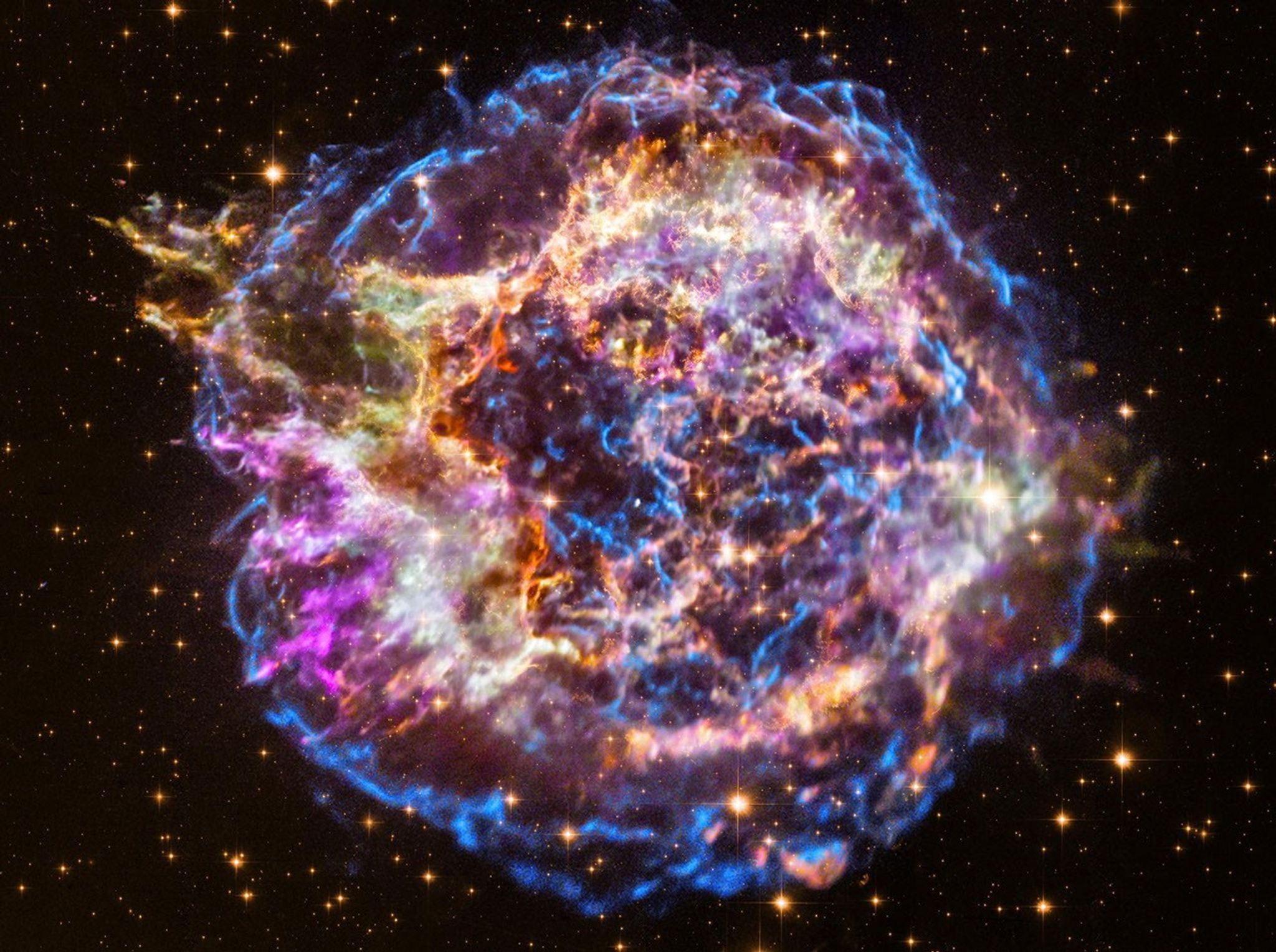 Colorful composite image of the Cassiopeia A supernova remnant, showing bright filaments of blue, purple, orange, and yellow gas and dust against a star-filled black background.