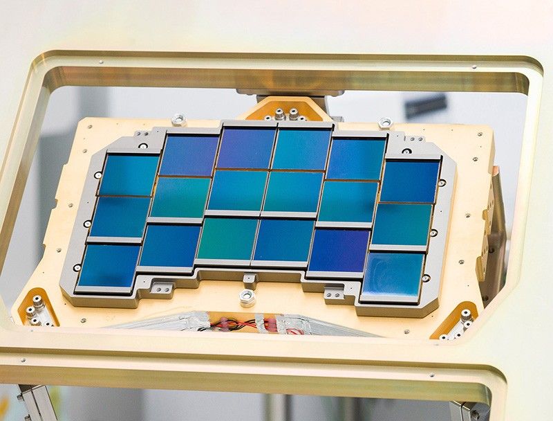 At the center of the photo is a set of 18 square detectors on a gold platform. They're arranged in six columns of three detectors such that the top of the middle two columns are close to the top of the image, the next two columns are a little lower down, and the two outer columns are even lower. The detectors are different shades of blue from royal blue to teal.