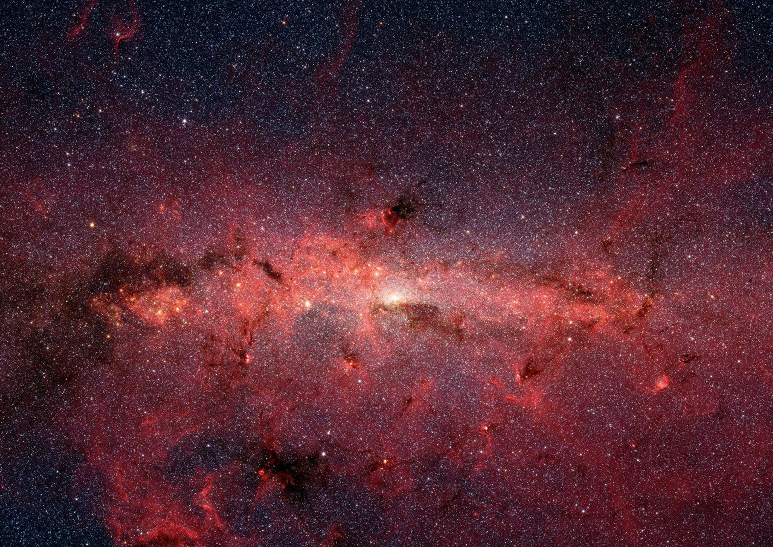 At the center of this starry vista is a bright white spot surrounded by warm knots of red clouds. The clouds extend across the image from left to right and nearly all the way from the bottom to the top, with some black sky visible toward the top of the image. Within the red wispy clouds, there are clouds that appear black, regions that are so dense, they are blocking our view.
