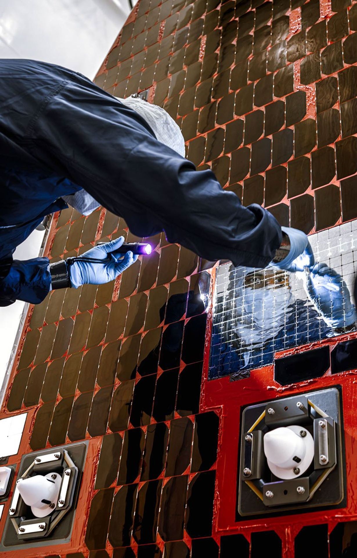 A technician wearing clean room garments and blue gloves inspects a square panel of shiny thermal tiles on NASA's Nancy Grace Roman Space Telescope, using a flashlight to check the surface. The reflective tile contrasts with the surrounding darker tiles and nearby white sensor housings.