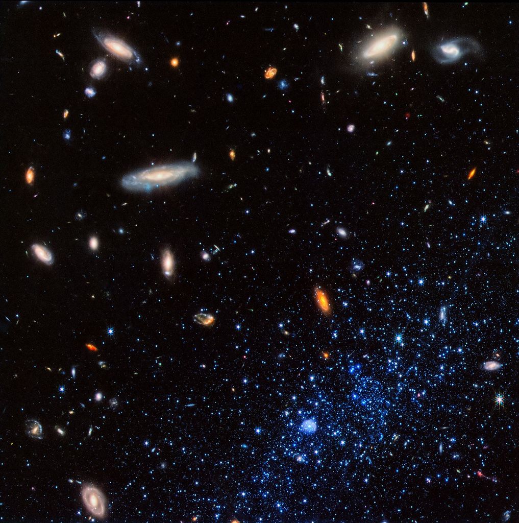 A concentration of bright blue stars occupies the bottom right corner of the image. At bottom center within them is a small blue bubble. The stars and bubble are part of a diffuse dwarf galaxy that extends beyond the image border. Background galaxies are scattered across the image, with some particularly prominent spirals located at upper left and upper right. The background of space is black.