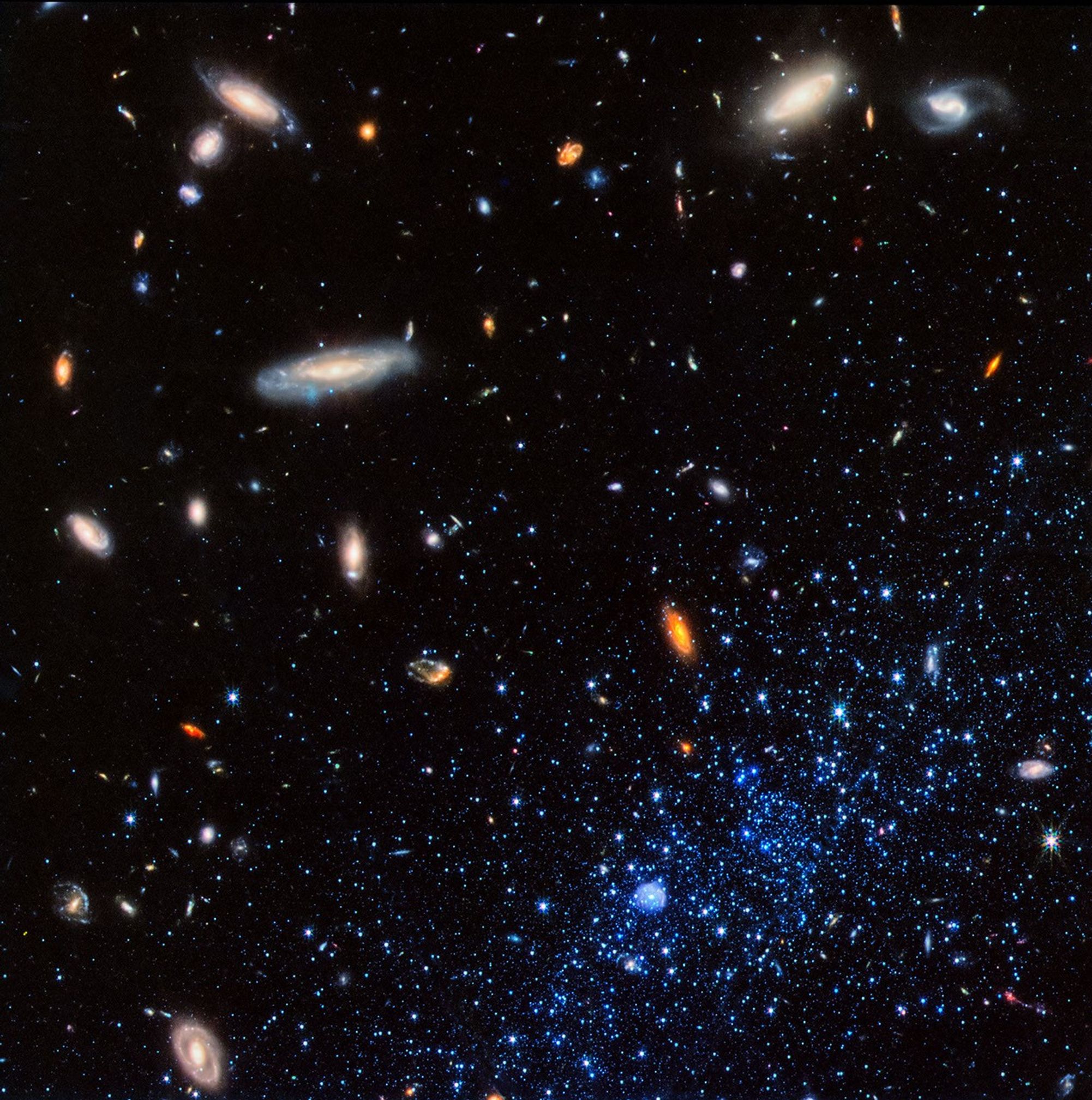 A concentration of bright blue stars occupies the bottom right corner of the image. At bottom center within them is a small blue bubble. The stars and bubble are part of a diffuse dwarf galaxy that extends beyond the image border. Background galaxies are scattered across the image, with some particularly prominent spirals located at upper left and upper right. The background of space is black.
