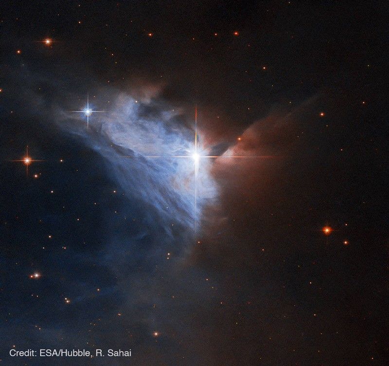 Near the center of the image shines a bright star with four spikes. It forms a vertex of a triangle of wispy blue and blue-white light that forms the emission nebula taking up about a third of the image. To the right and above the bright star, the image quickly fades from pale red to black, which is where a dense cloud of dust blocks our view of the nebula. Credit: ESA/Hubble, R. Sahai