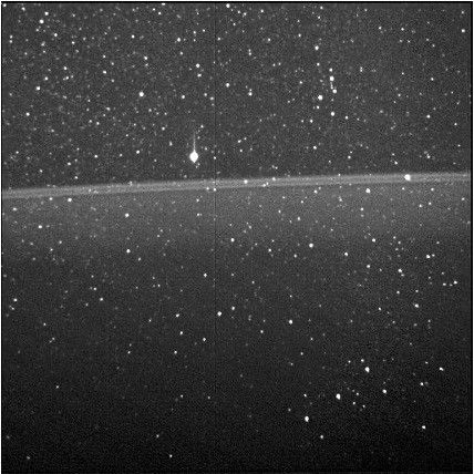 A double line of light gray breaks up a starfield dotted with points of light, running from left to right about a third of the way down this image. The image is looking through Jupiter’s main ring. The star field’s background is darker at the top and bottom of the image, with a gradient of gray from lighter to darker running below the double line.