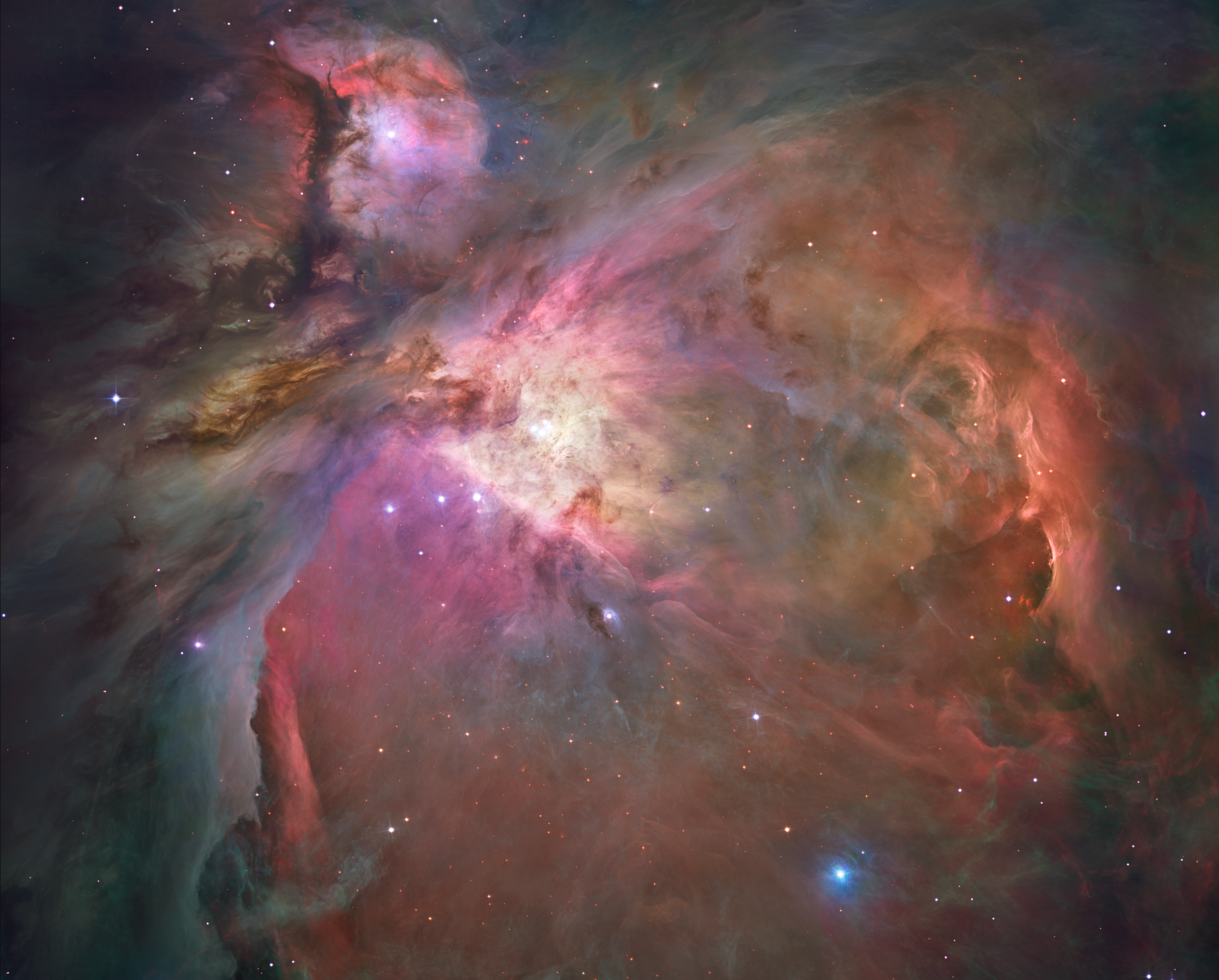 This Hubble image of the Orion Nebula offers a peek inside a cavern of roiling dust and gas where thousands of stars are forming. The image is dominated by wispy clouds in shades of red, yellow and orange against a dark background and dotted with stars. There are two concentrations of clouds, one in the shape of an anatomical heart anchored at the center of the image and tilted so that it fills the right side and lower left. In the upper left, separated from the heart shape with a dark lane is a knot of reds and pinks.