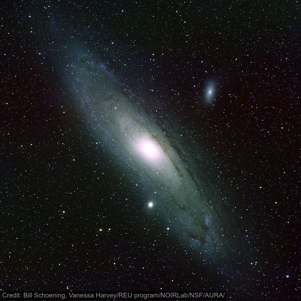 The background of this image is dotted with small circles of light in white and shades of pale yellows, reds, or blues. In the center of the image is a large, elongated oval angled from the top left to bottom right with is the disk of the Andromeda galaxy. At the center of the disk is a circle of bright white, with the rest of the disk filled by hazy white. There is also a smaller bright oval above and to the right of the center of Andromeda, which is one of its companion galaxies. The image is watermarked: “Credit: Bill Schoening, Vanessa Harvey/REU program/NOIRLab/NSF/AURA.”