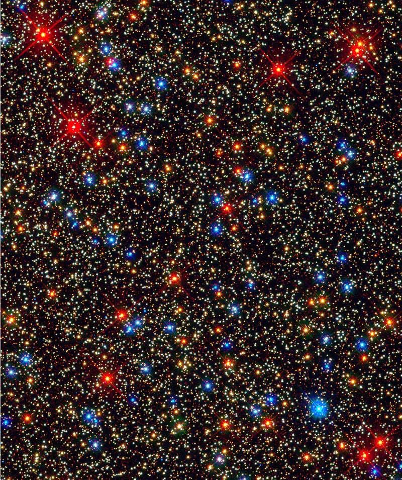 A black background is speckled by a seemingly infinite number of bright spots, with smaller spots being a pale-yellow color with some brighter and larger spots that are orange, bright red, and blue.