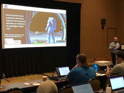 Presenter stands near a projection screen showing a slide on mitochondrial metabolic alterations and spaceflight health risk, as attendees watch and take notes on laptops.