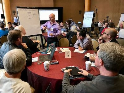 Facilitator speaks to a small group seated around a round table during a workshop session, with notes on a flip chart and a presentation screen in the background.