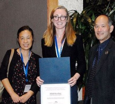 Three people pose for a photo, with the center person holding an award certificate and wearing a medal.