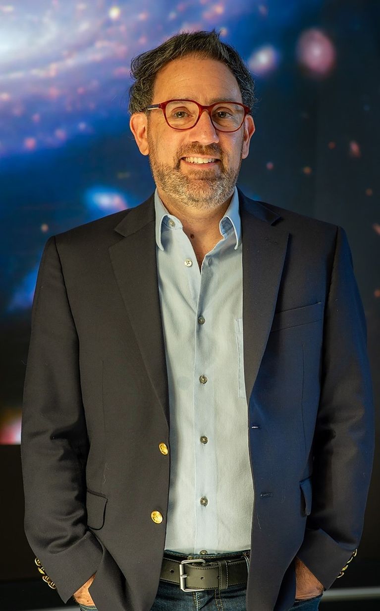 Male, dark hair, peppered beard with red frame glasses, standing with hands in pockets, wearing a dark blue blazer and light blue button-down shirt.