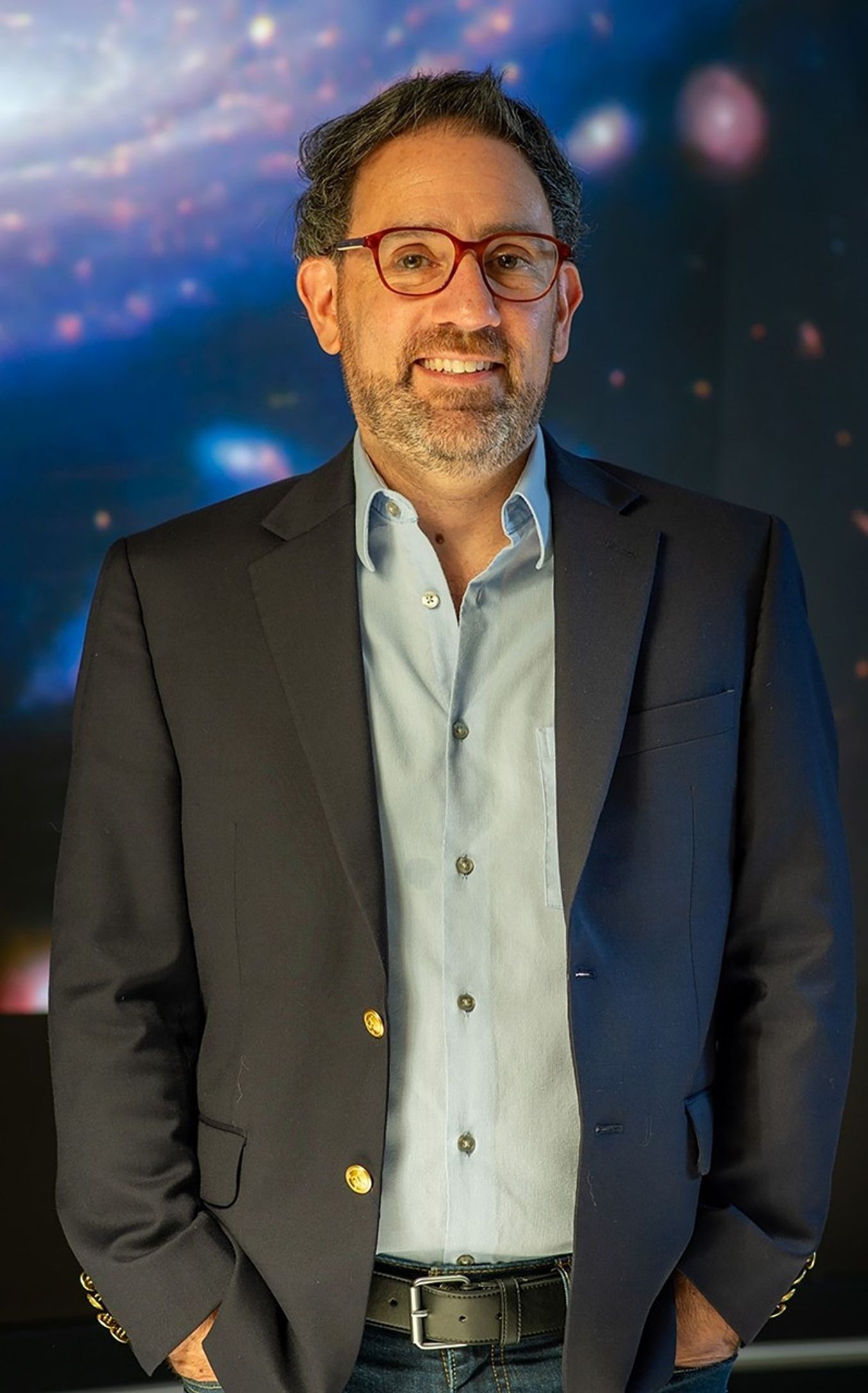 Male, dark hair, peppered beard with red frame glasses, standing with hands in pockets, wearing a dark blue blazer and light blue button-down shirt.