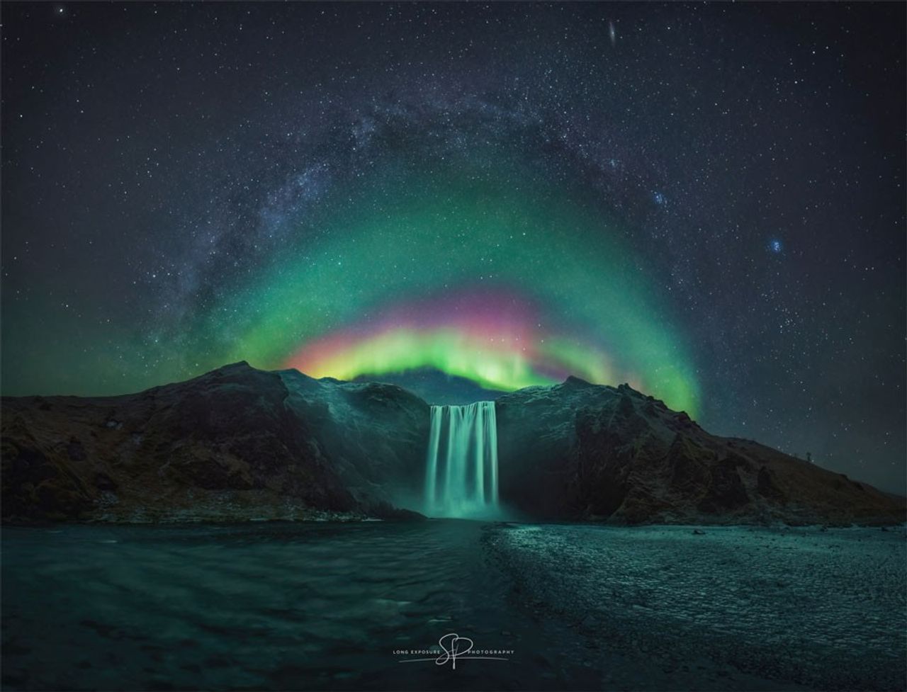 A waterfall is shown in the image center below a starry sky. Arching above the waterfall is a colorful aurora. Arching above the aurora is the central band of the Milky Way. Please see the explanation for more detailed information.