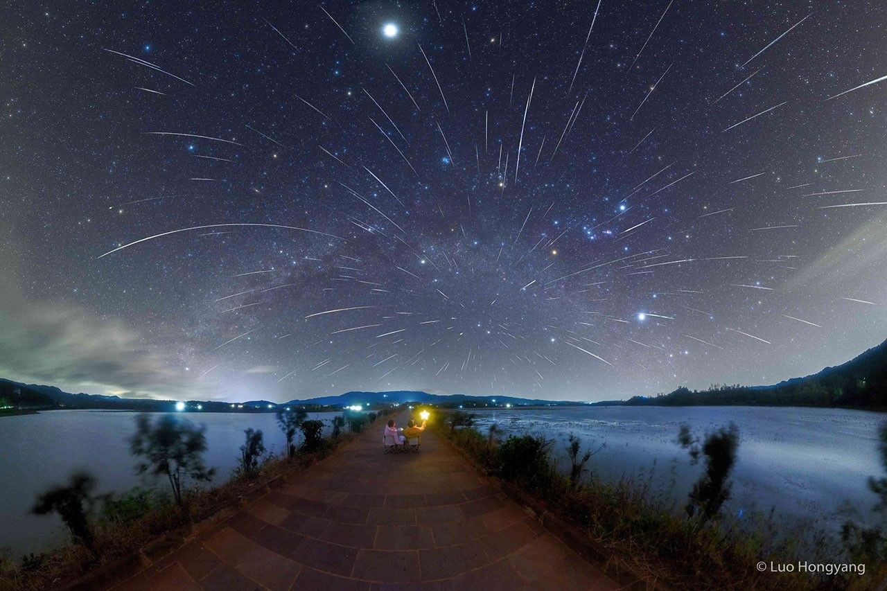 Two people are pictured from the back looking at a dark star-filled sky. The sky is also filled with numerous streaks caused by meteors from the Geminids meteor shower. Please see the explanation for more detailed information.