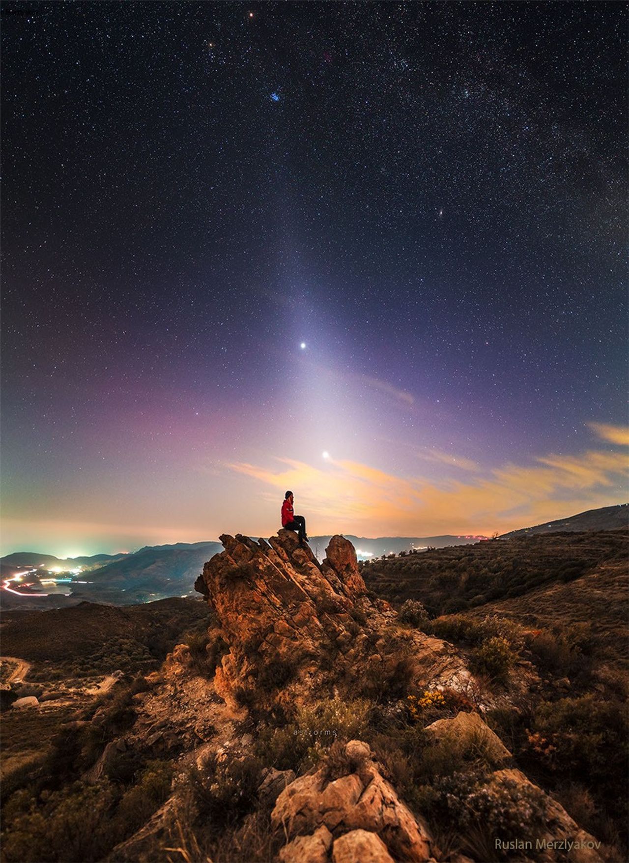 A person is seen sitting on a rock under an unusual sky. In the sky above is light diffuse band extending down to the horizon that goes through two bright dots, Jupiter and Venus. The Pleiades star cluster is visible above them. Please see 