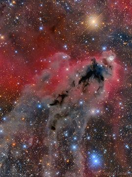 An image of a foreboding dark nebula before a red-glowing gas background and many bright and colorful stars. Please see the explanation for more detailed information.