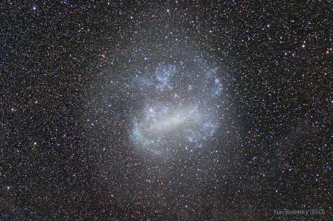 A galaxy is pictured that appears mostly blue and white with a prominent bar across its center. The galaxy is the LMC, and thousands of dim stars from our Milky Way, in the foreground, complete the frame. Please see the explanation for more