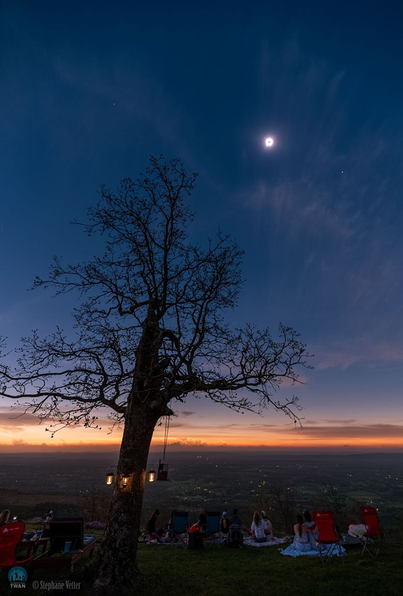 A totally eclipsed Sun is seen in the sky surrounded by a bright corona. In the foreground several people watch it near a large tree. To the right of the eclipsed Sun is the bright planet Venus, while the nearly-as- bright planet Jupiter is