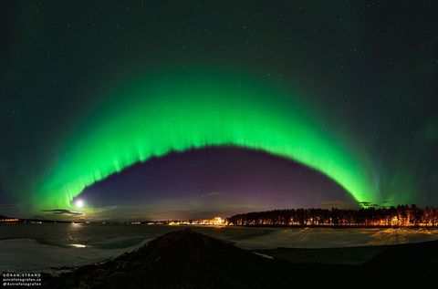A big green arc is seen arching across the night sky. The arc fades away above into a green haze, while no green glow is seen below the arc. A dark sky filled with stars and constellations fills the background. Snow and distant trees line t
