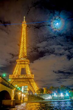 The famous Eiffel Tower in Paris, France is pictured on the left lit up in gold at night. A blue laser shines out from the top. Clouds dot the background sky. The Moon is also visible through the clouds, but is circled by colorful rings: a 