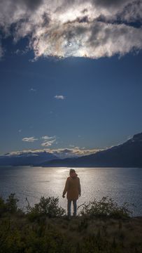 A person stands looking over a lake. High in a partly cloudy sky is the Sun. A close look at the Sun will show that there is a dark spot in the center -- the Moon during an annular eclipse. Please see the explanation for more detailed infor