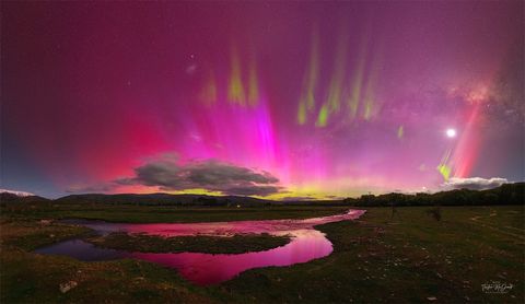 A night sky is shown that appears mostly red due to pervasive aurora. In the foreground is covered by watery grasslands. Clouds are visible above the horizon. Thin green aurora are visible toward the top of the frame. In the background one 
