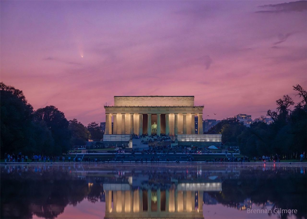The Lincoln Memorial monument in Washington, DC, USA is pictured from afar. Behind the monument is a sunset-colored pink sky. In the sky, on the upper left, is a white streak that is a comet. Please see the explanation for more detailed inf