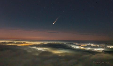 A picture shows clouds across the bottom and a dark night sky across the top. In the middle is a band of orange sky. City lights are visible on the right through gaps in the clouds. In the center of the upper sky is a comet with its tail po