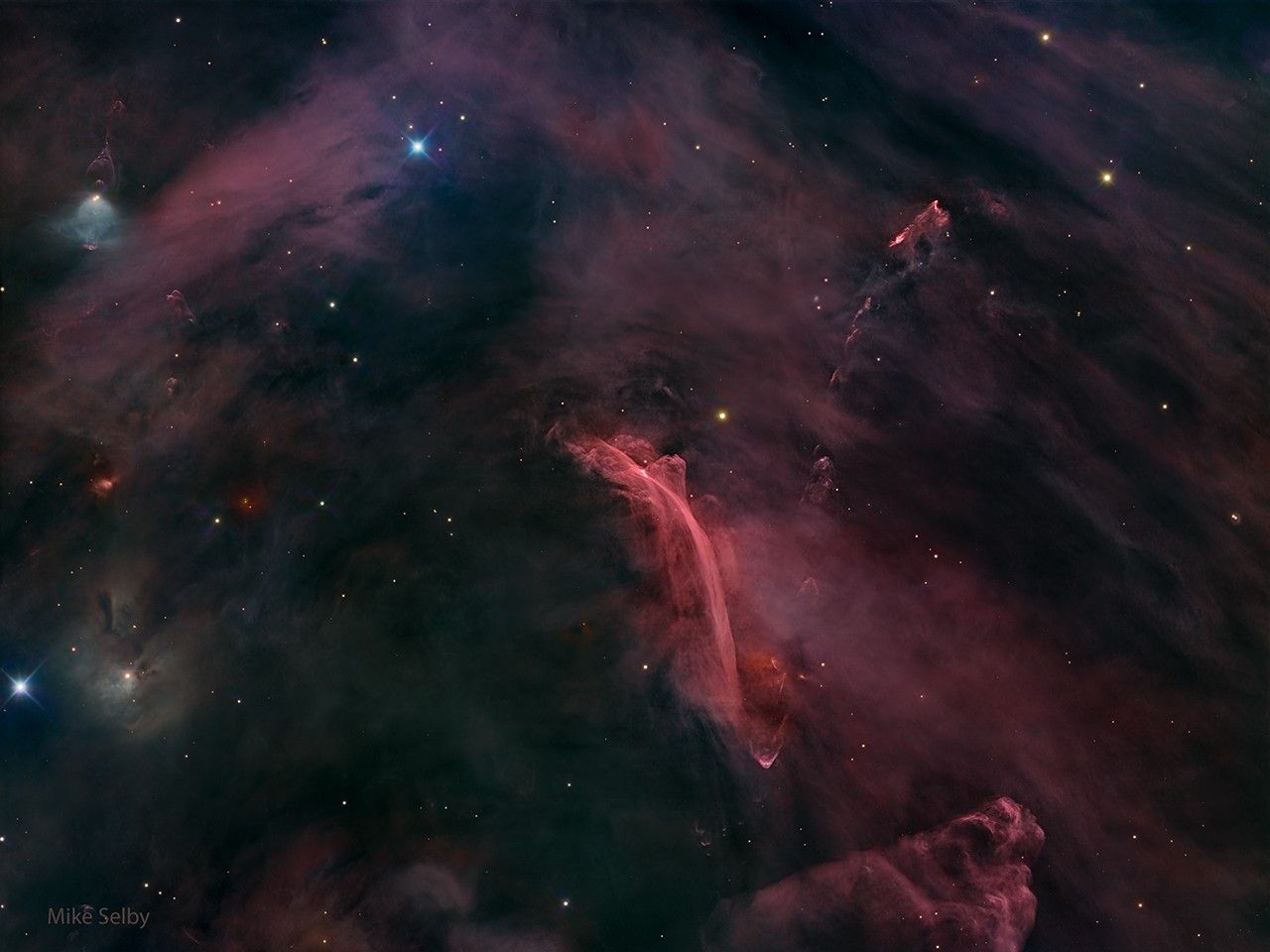 A starfield filled with a diffuse red glow has an unusual nebula on the lower left. The nebula has bright red filaments that curve down and appear to be reminiscent of a waterfall on Earth. Please see the explanation for more detailed infor