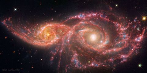 Two spiral galaxies are pictured on the left and right. They galaxy on the left is smaller. Both show red lanes of dust in their spiral arms. Please see the explanation for more detailed information.