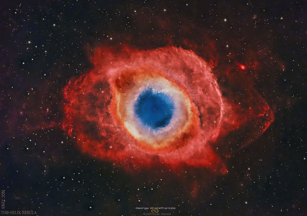 A nearly circular nebula with a blue core surrounded by small white knots, an orange ring and expansive red strucures. Please see the explanation for more detailed information.