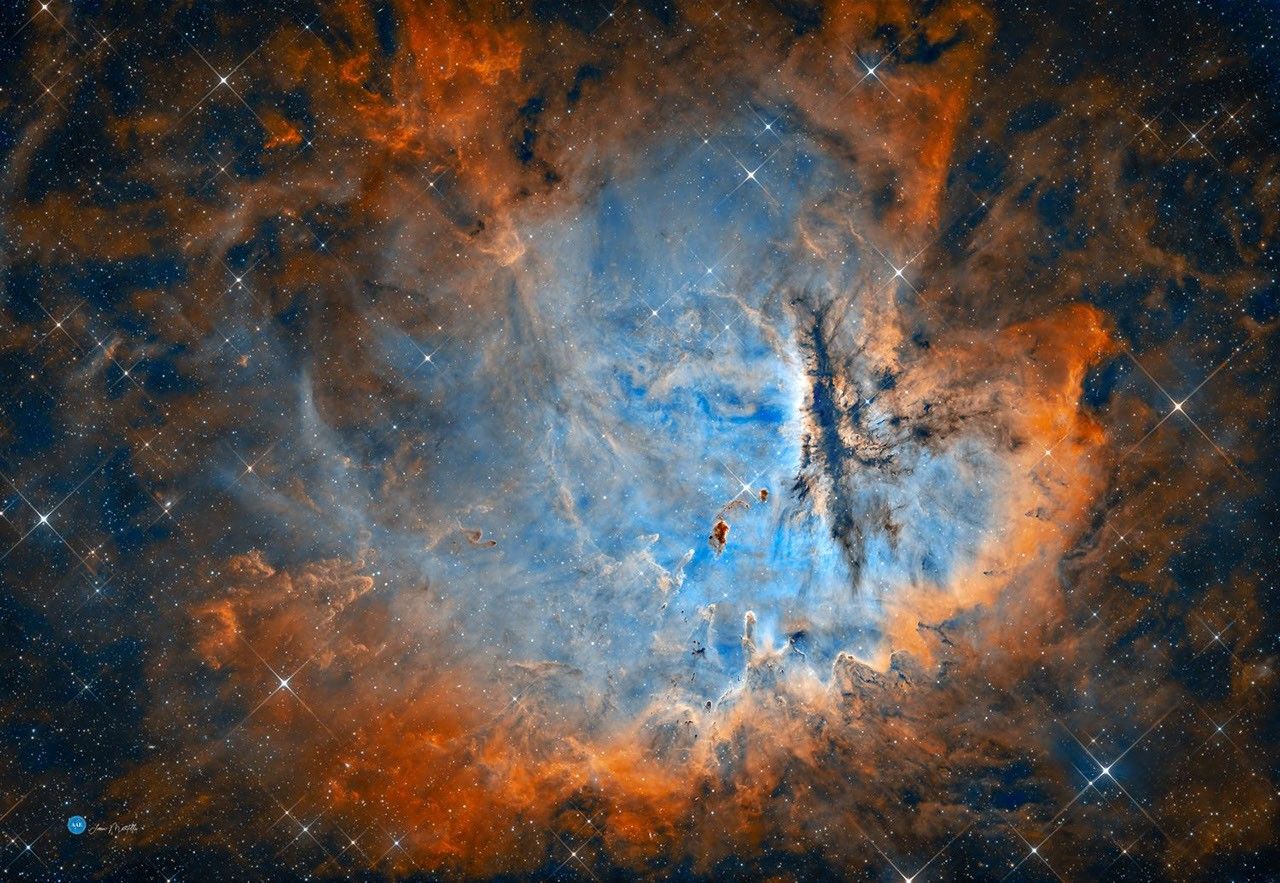 A starfield dominated by a large nebula is pictured. The center is blue and the perimeter is red. Many dark dust pillars are visible. Please see the explanation for more detailed information.
