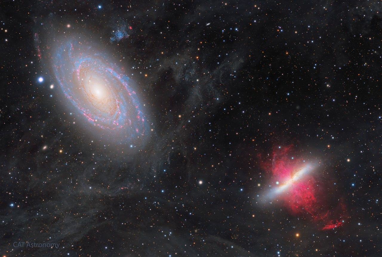 Two large galaxies are shown against a dark starfield. The galaxy on the upper left has blue spiral arms speckled with red nebulae. The galaxy on the lower right has a white line with red filaments on each side. Thin wisps cover some of the