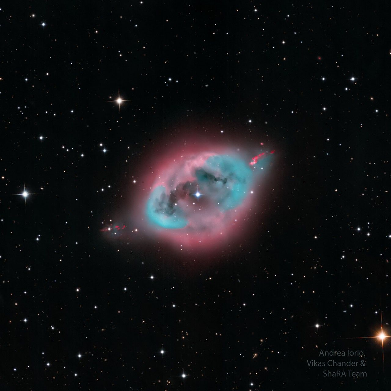 A dark starfield highlights a blue and pink nebula in its center. Some dark lanes of dust are seen inside nebula's center. Please see the explanation for more detailed information.
