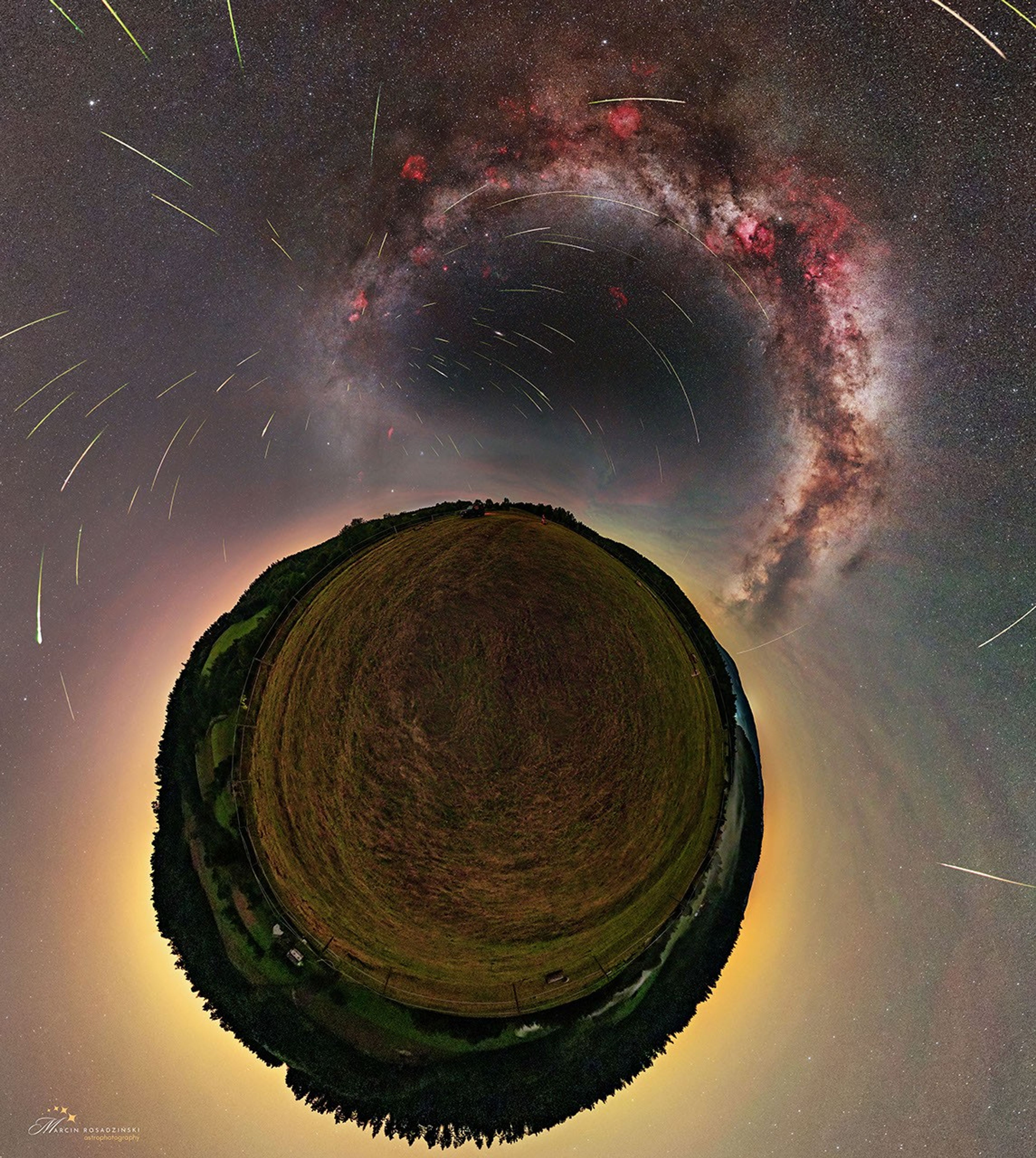 The ground is shown in a little planet perspective, surrounded by the sky with a diffuse glow. The Milky Way is visible in the sky, as well as some nebulae in red and over a hundred meteors. Most of them appear to come from the same point.
