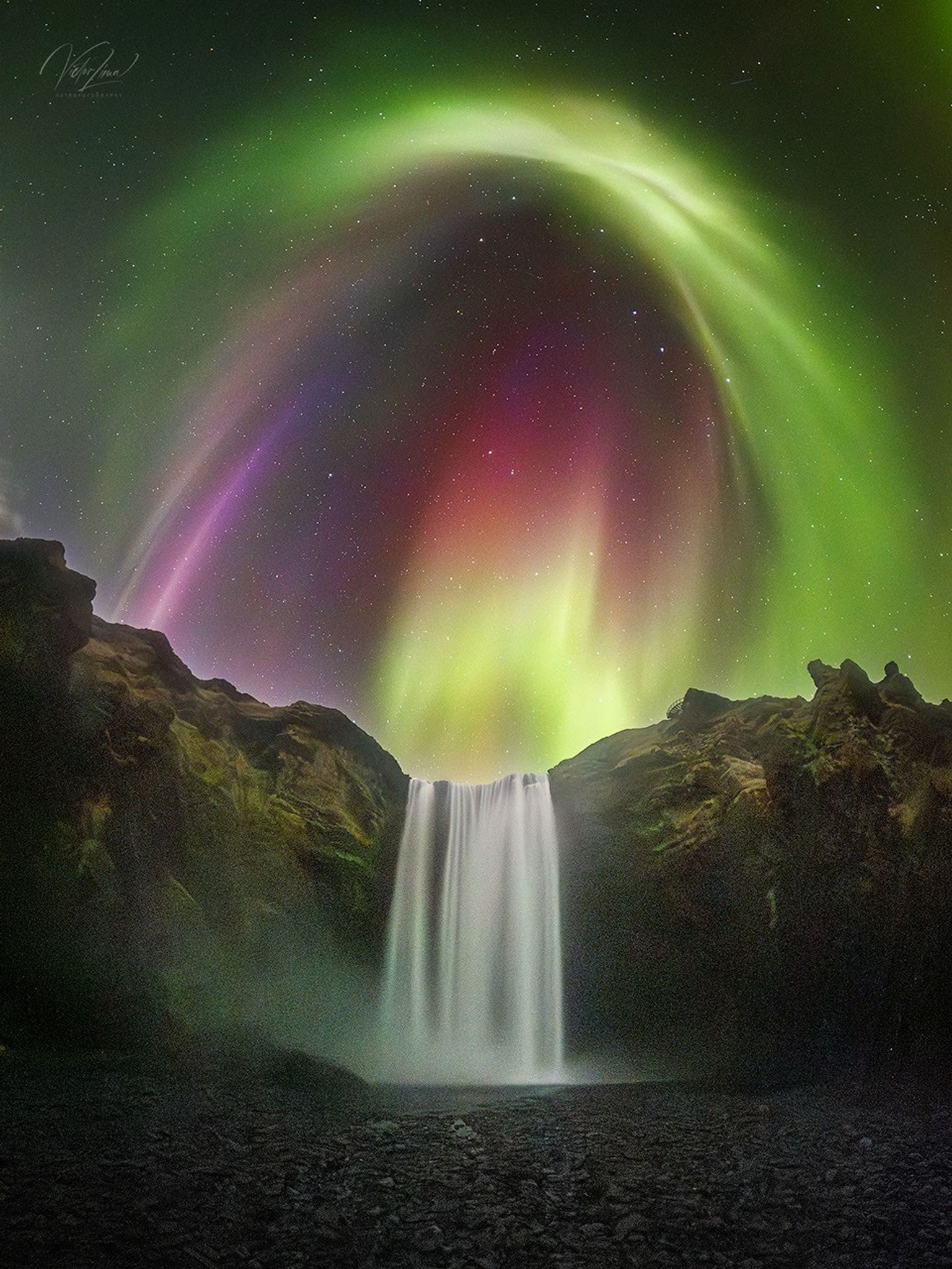 Aurora with green, pink and yellow shades over a waterfall will stars in the background.