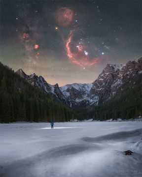 A person stands in the distance on a snowy landscape shining a flashlight on the ground. Trees and snow capped mountains are on the horizon. In the starry sky above, several iconic red nebulas are visible. Please see the explanation for mor