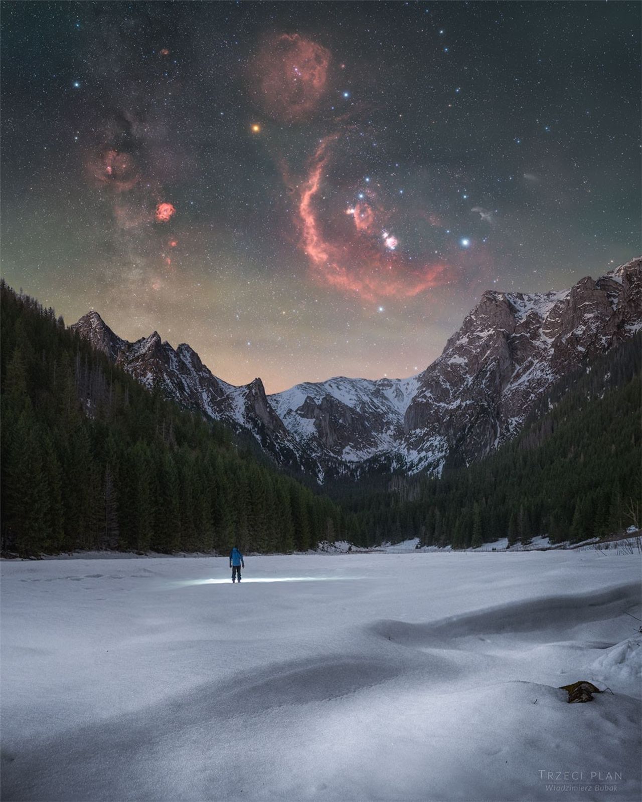A person stands in the distance on a snowy landscape shining a flashlight on the ground. Trees and snow capped mountains are on the horizon. In the starry sky above, several iconic red nebulas are visible. Please see the explanation for mor