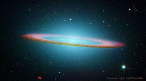 A red-tinged ring of dust is seen nearly on edge. In the ring's center and extending around the frame, blue gas and stars are shown. Please see the explanation for more detailed information.