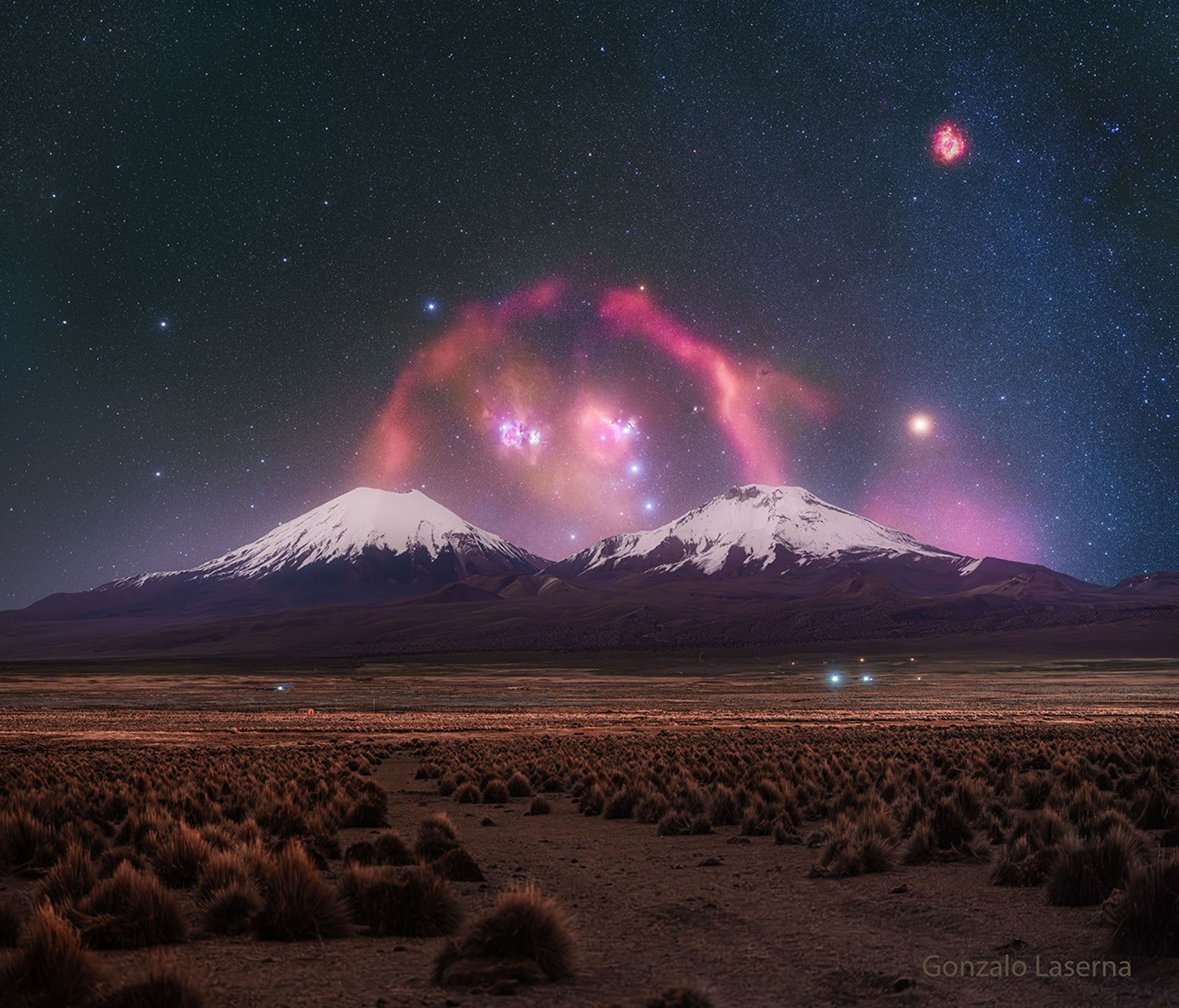 A pair of snow topped volcanoes are seen in the distance. A background starry sky is seen behind them, with a red filamentary nebula looping around that appears to connect the two tops of the volcanoes.