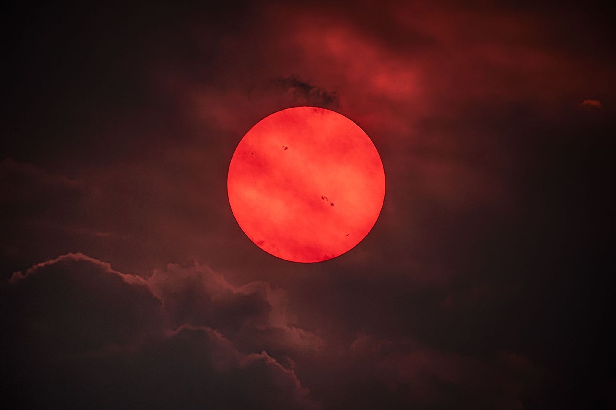 A deep red sun shows several sunspots. Clouds and haze are visible.