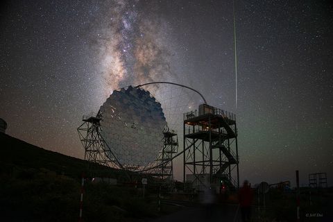 A large, segmented telescope mirror is seen against the night sky along with  the Milky Way and a bright meteor.