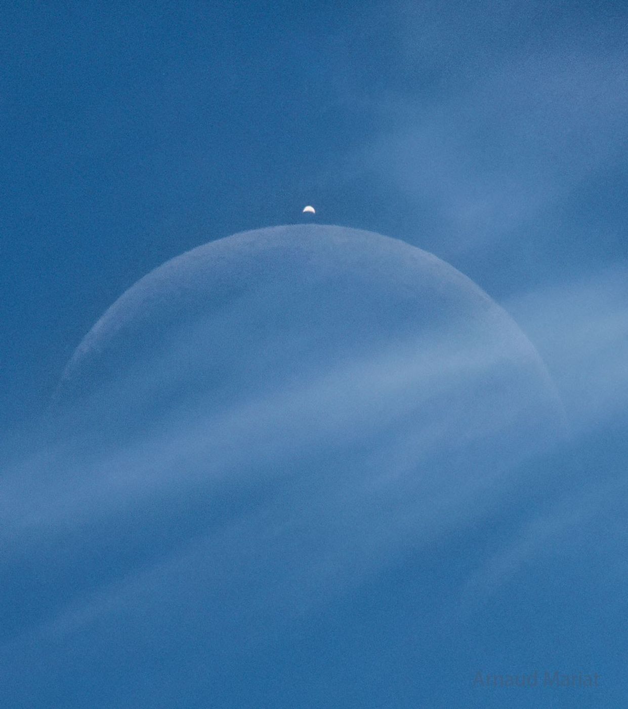 A blue sky has a light cloud crossing the middle of the image. Two crescent objects are visible through the cloud, a large faint crescent in the center and a brighter crescent above it.