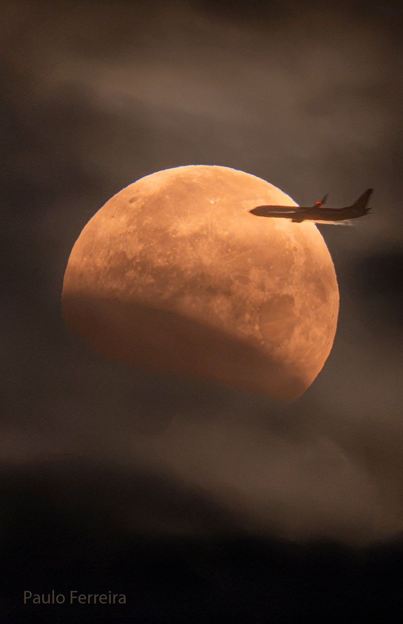 The Moon is seen in front of clouds but with part of its lower left being unusually dark. On the upper right, a foreground airplane appears flying into the disk of the Moon.