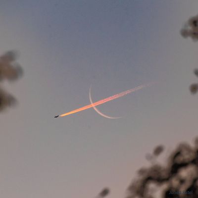 A thin crescent moon is shown with a bright red  contrail going through it, right to left.  Please see the explanation for more detailed information.